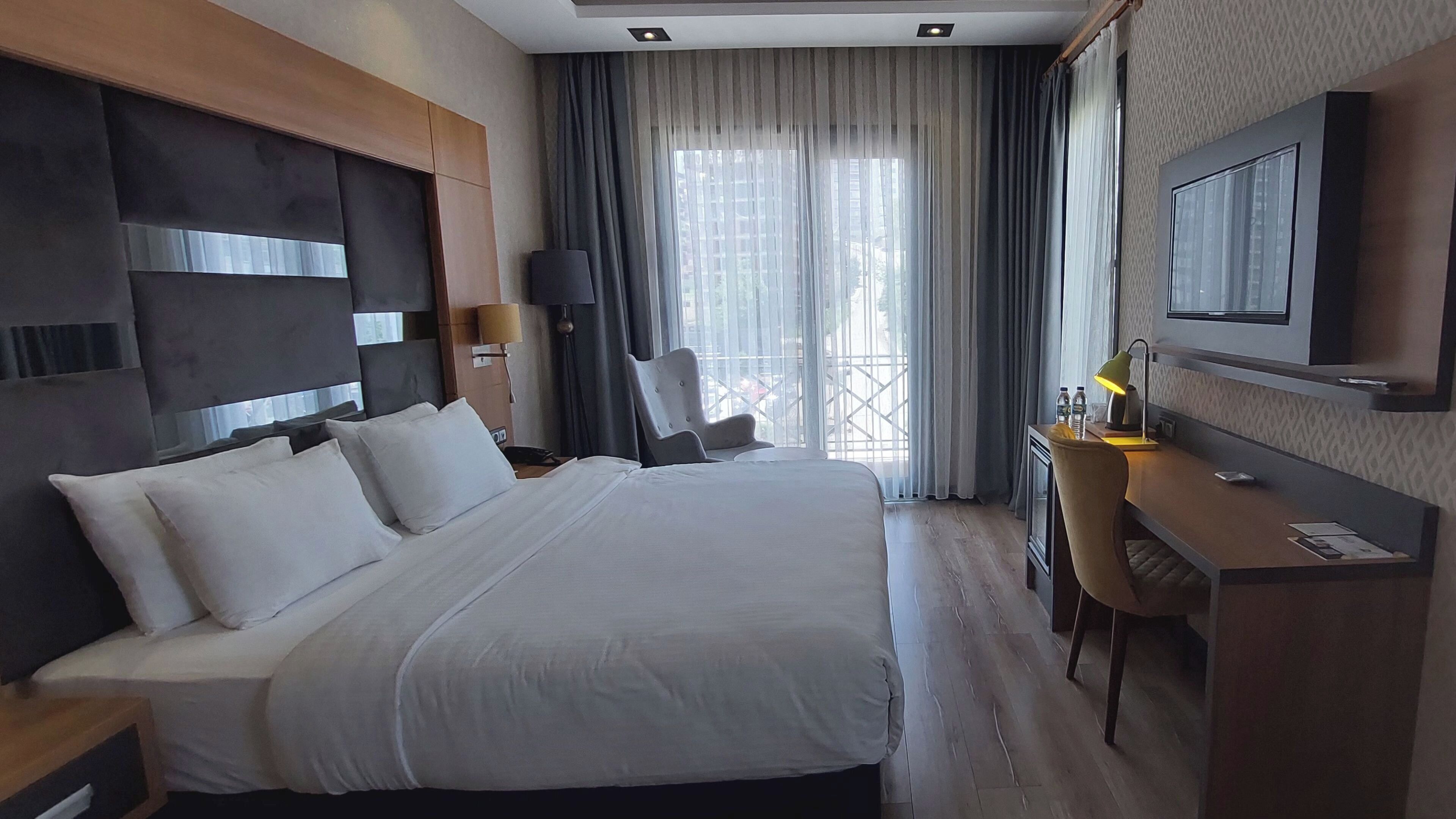 A hotel room with a large bed, a desk, a chair, a TV, and a window with curtains.