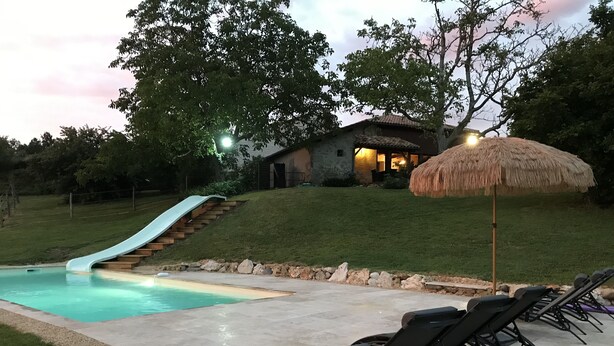 A backyard with a swimming pool, a slide, lounge chairs, and a thatched umbrella.
