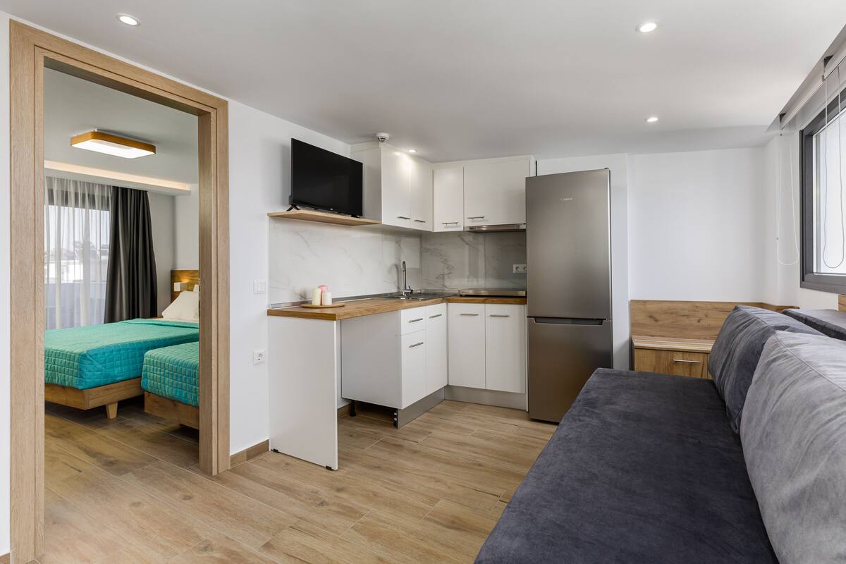 Una habitación compacta y moderna con kitchenette, una cama con ropa de cama verde y un sofá gris.