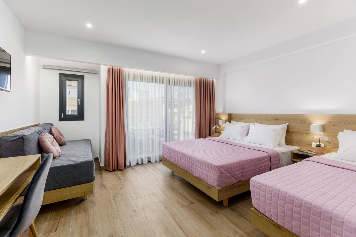 Habitación de hotel con dos camas, un escritorio, una silla y un ventanal con cortinas translúcidas.