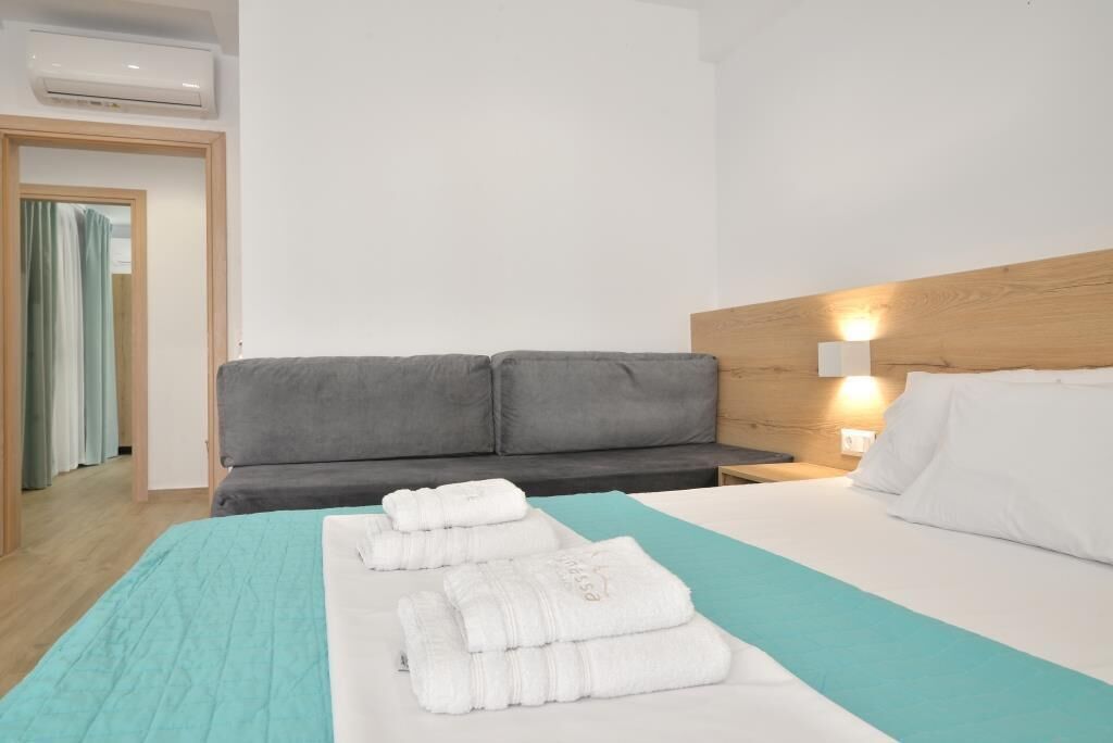 Et hotelværelse med en grå sofa, en seng med hvidt og teal sengetøj samt en vægmonteret lampe.