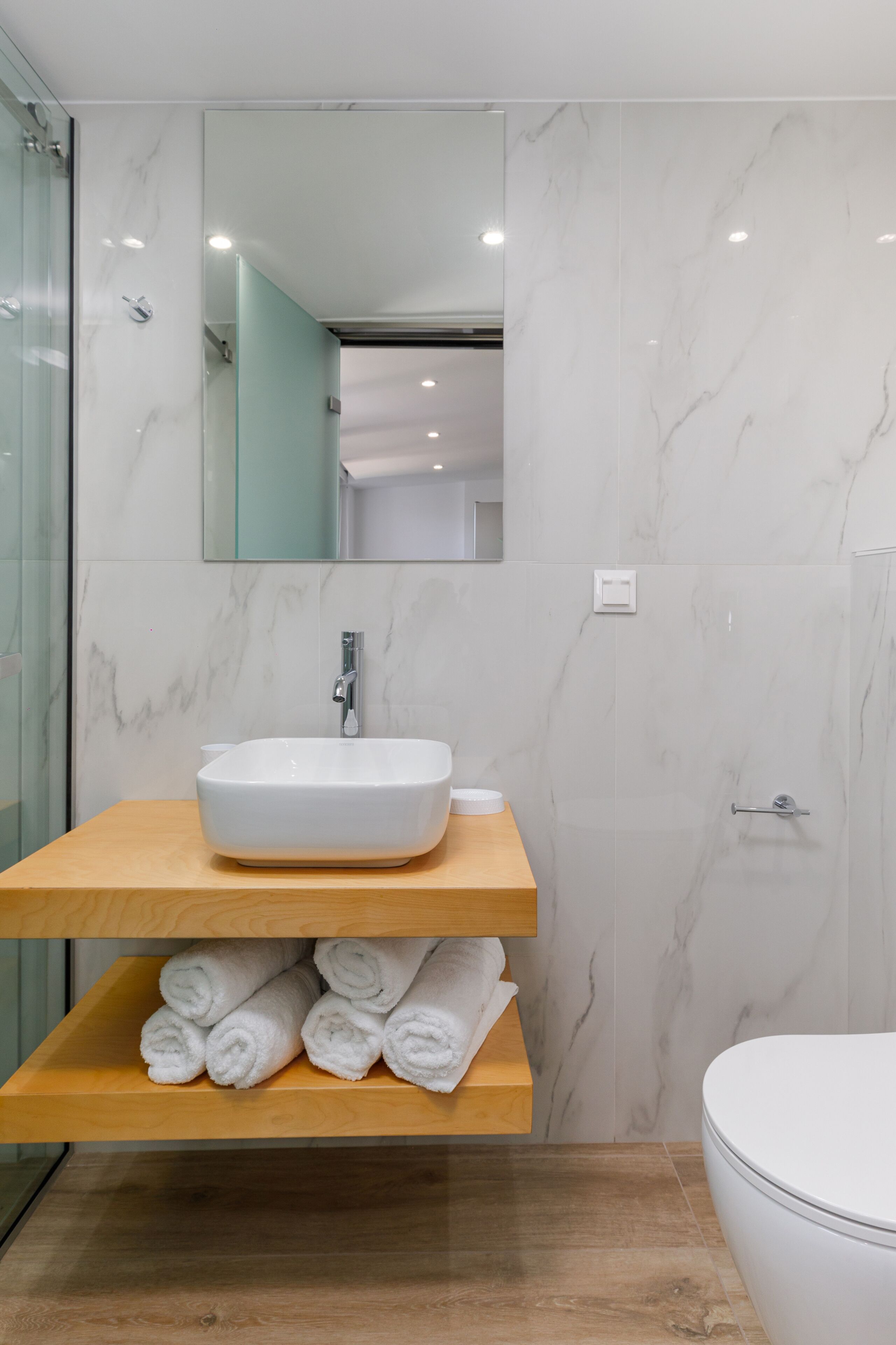 Un baño moderno con pared de mármol, tocador de madera, lavamanos blanco y un inodoro.