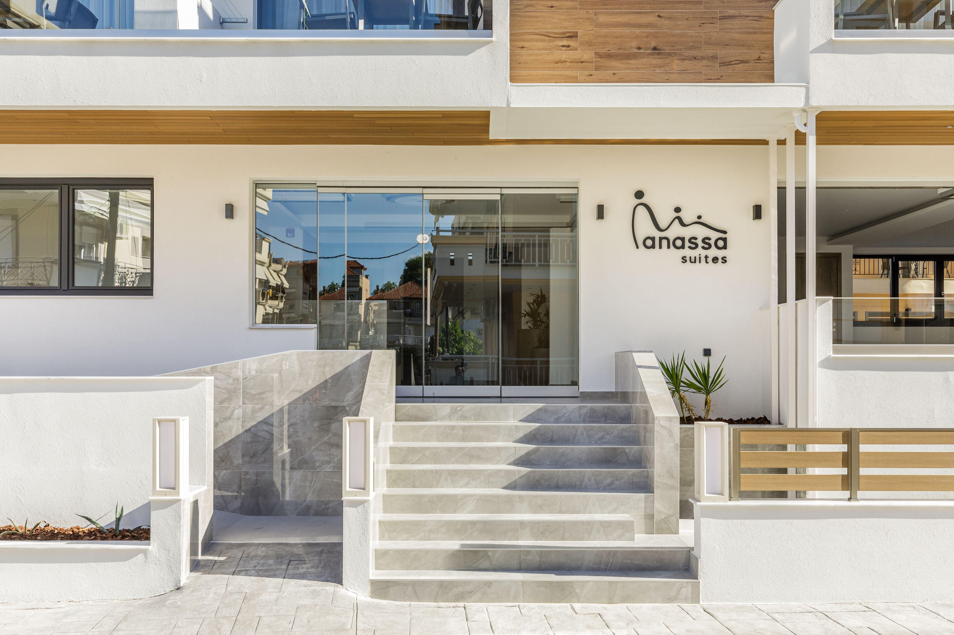 Um edifício moderno com entrada de vidro, um banco de madeira e uma placa com 'Anassa Suites'.