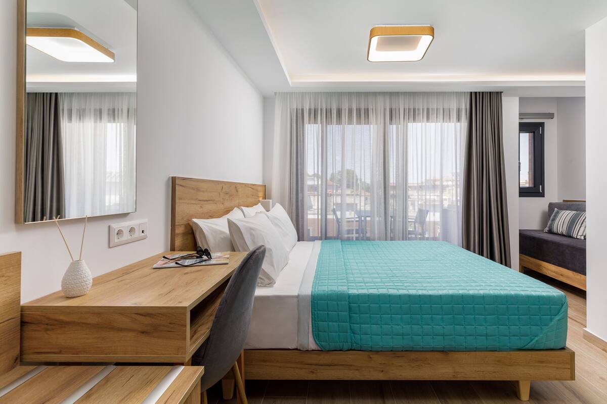 Habitación de hotel con cama de madera, un escritorio y una silla. Hay una ventana con cortinas, un espejo y vistas a edificios.
