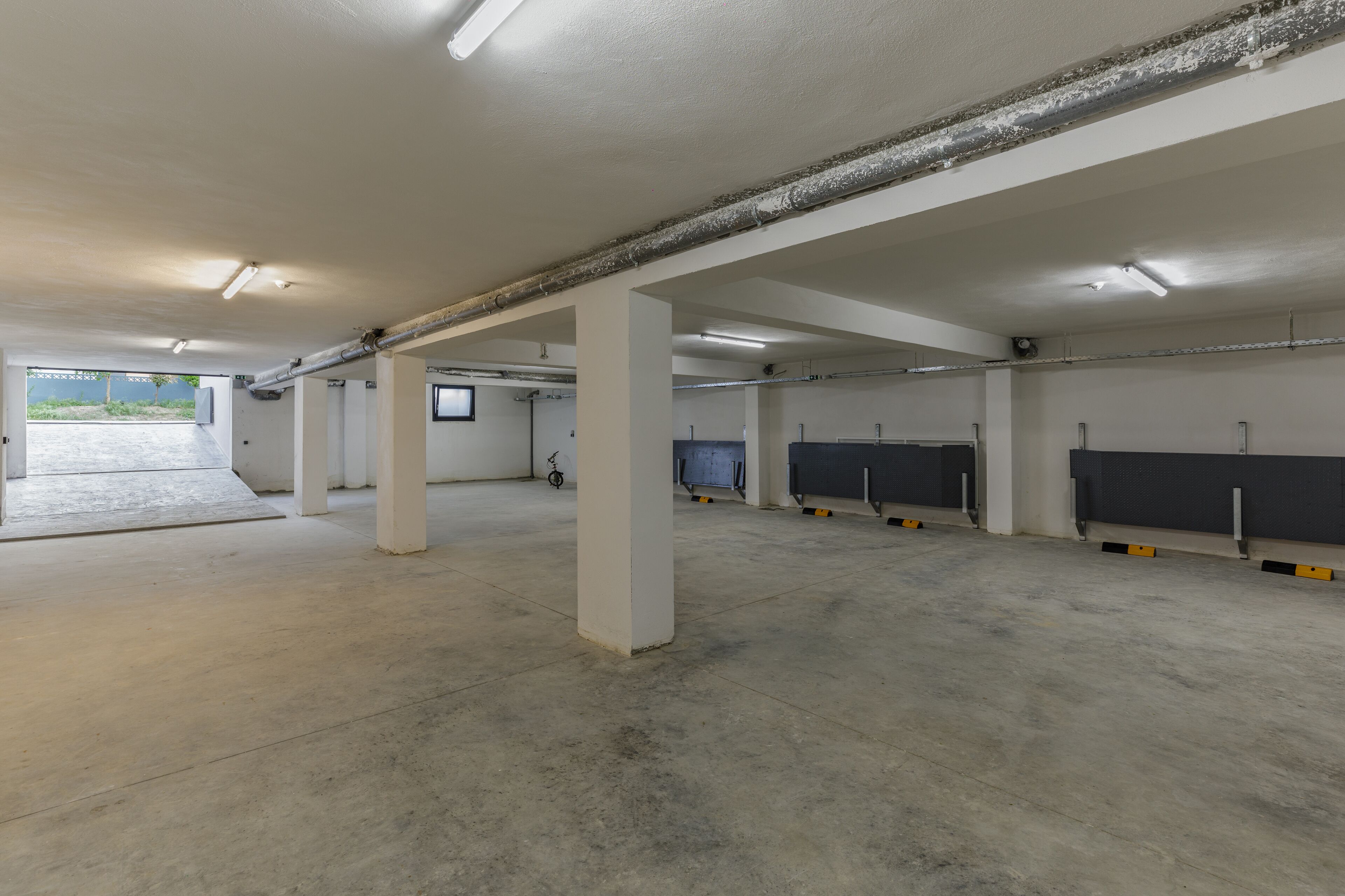A spacious indoor parking area with concrete floors and white pillars, featuring a few bicycles and a small TV screen.