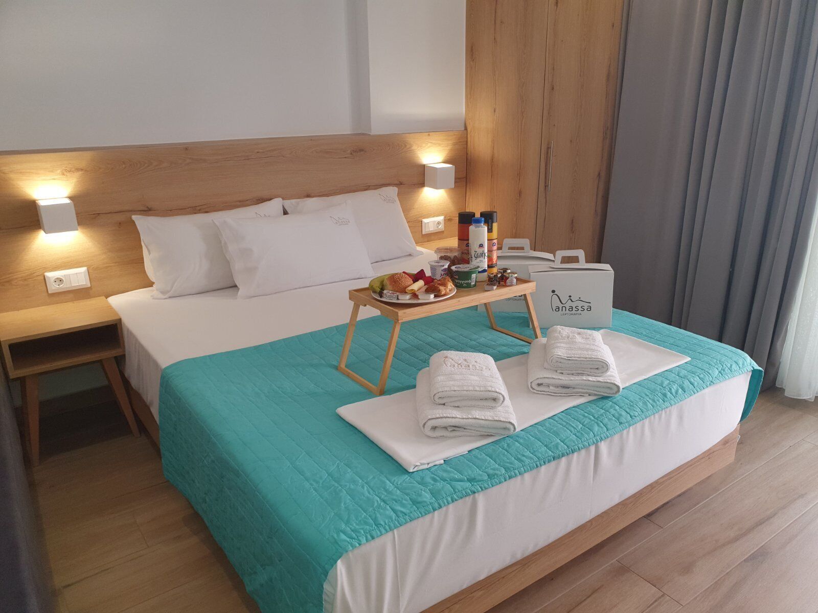 A hotel room with a bed, a nightstand, a tray table with breakfast items, and a sign for 'Anassa LE PIKOTARIA'.