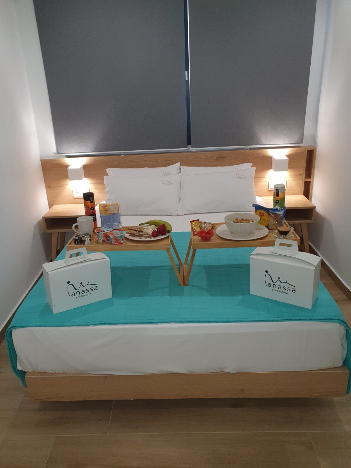 Una cama bien hecha con una colcha color turquesa, dos mesitas de noche con lámparas, una bandeja con alimentos para el desayuno y dos cajas blancas con el logotipo 'anassa'.