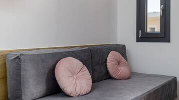 A grey sofa with wooden legs and two pink cushions, positioned against a white wall with a mounted TV and a window with a black frame.