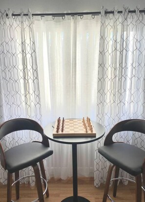 Um tabuleiro de xadrez sobre uma pequena mesa redonda com duas cadeiras, com um pano de cortina estampado ao fundo.