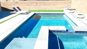 Et rektangulært swimmingpool med to afdelinger, hver med forskellig dybde. Liggestole og en lille terrassemed ildsted.