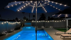 Une piscine avec un parasol, des chaises longues et des guirlandes lumineuses.