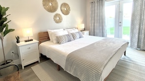 Une chambre à coucher avec un grand lit, deux tables de chevet, une plante en pot, des œuvres d’art décoratives au mur et des rideaux à motifs.