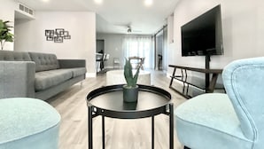 Un salon moderne avec un canapé gris, un fauteuil bleu, une table basse ronde noire agrémentée d’une plante, et un téléviseur fixé au mur.