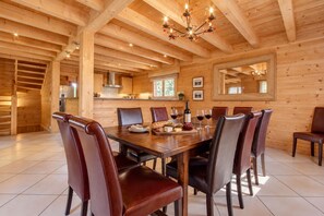 A spacious dining area with a wooden table and chairs, a chandelier, and a kitchen in the background.