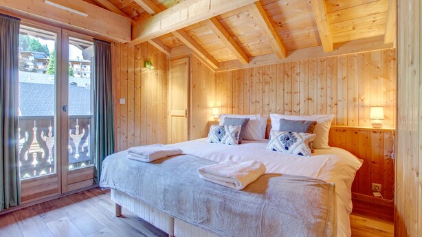 A wooden-paneled bedroom with a large bed, two bedside lamps, and a view through a sliding glass door. - Alptitude Chalet Largo - spacious detached chalet (Morzine)