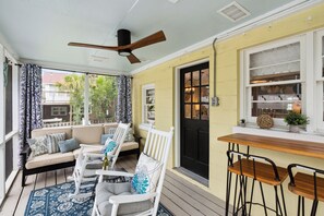 A sunlit porch with a wooden table, white bar stools, a cushioned seating area, and a ceiling fan. - Pleasant Breeze | Steps to Beach, Screened Porch, Fenced Yard + Pet Considered (Folly Beach)