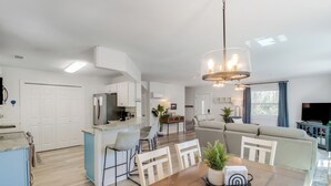 Une cuisine moderne avec des meubles blancs, un coin repas avec une table et des chaises en bois, un salon avec un canapé et une télévision, et un lustre.