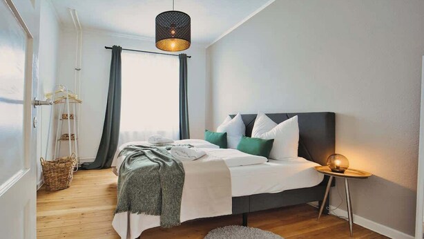 Un dormitorio con cama, una lámpara de noche, una ventana con cortinas, una lámpara colgante y una alfombra redonda pequeña.