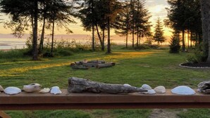 Un banc en bois orné de coquillages, une bûche et un foyer, le tout installé dans un espace verdoyant, agrémenté d’arbres et offrant une vue sur le coucher de soleil.