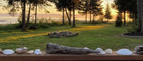 Un banc en bois orné de coquillages, une bûche et un foyer, le tout installé dans un espace verdoyant, agrémenté d’arbres et offrant une vue sur le coucher de soleil.