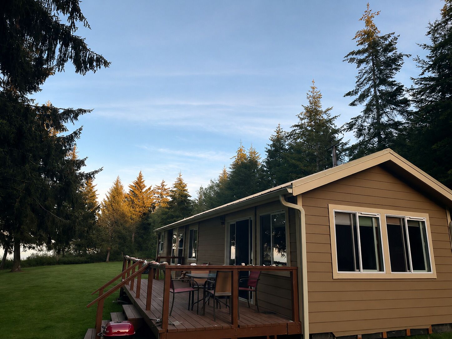 A single-story house with a deck, large windows, and a grassy yard surrounded by trees.