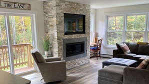 A living room with a stone fireplace, a flat-screen TV, and a large window overlooking a deck.