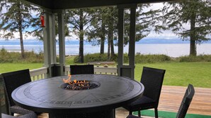 A covered outdoor seating area with a circular table and chairs, a fire pit, and a view of a lake and trees. - Beachfront House- The Sea Eagle (Sekiu)