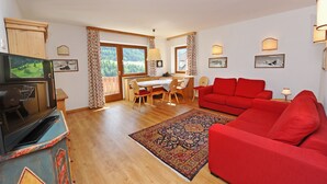 A living room with a red sofa, a dining table with chairs, a television, and a large window with curtains. - Apartments Valverda 111A (Ortisei)