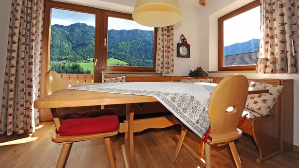 A cozy room with a wooden table and chairs, large windows with a mountain view, and a hanging lamp. - Apartments Valverda 111A (Ortisei)