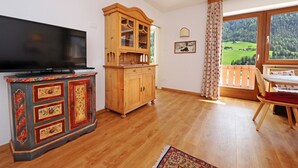 A room with a wooden cabinet, a television, a dining table with chairs, and a large window with a view of a green landscape. - Apartments Valverda 111A (Ortisei)