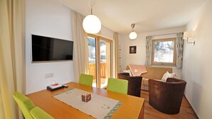 Uma sala de jantar com uma mesa de madeira, cadeiras verdes, uma TV na parede e um sofá.