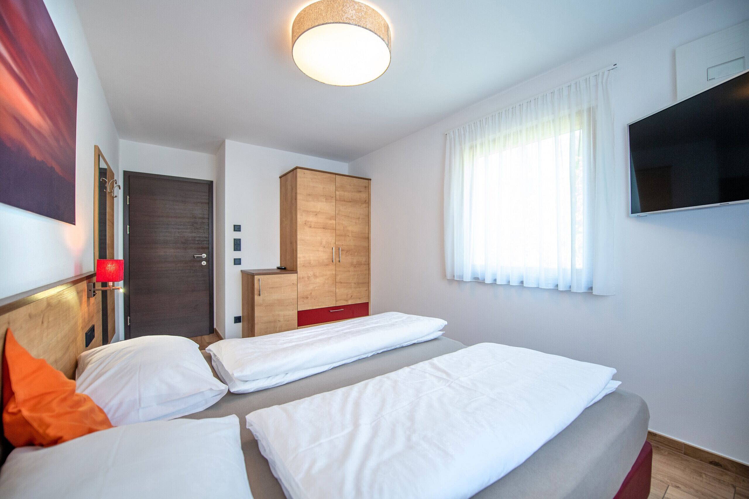 Hotellihuone, jossa on kaksi sänkyä, puinen vaatekaappi, televisio ja suuri ikkuna, jossa on verhot.