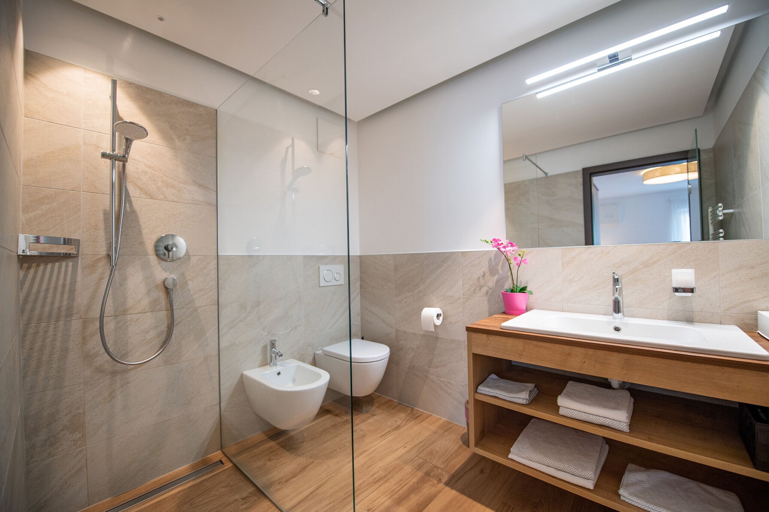 A modern bathroom with a glass-enclosed shower, a bidet, a toilet, and a wooden vanity with a sink and towel racks.