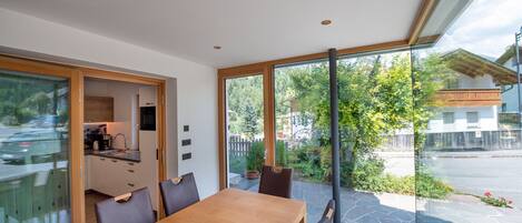 Un espace repas moderne avec une table et des chaises en bois, une cuisine en arrière-plan et une vue sur un jardin et une rue, le tout visible à travers de grandes fenêtres.