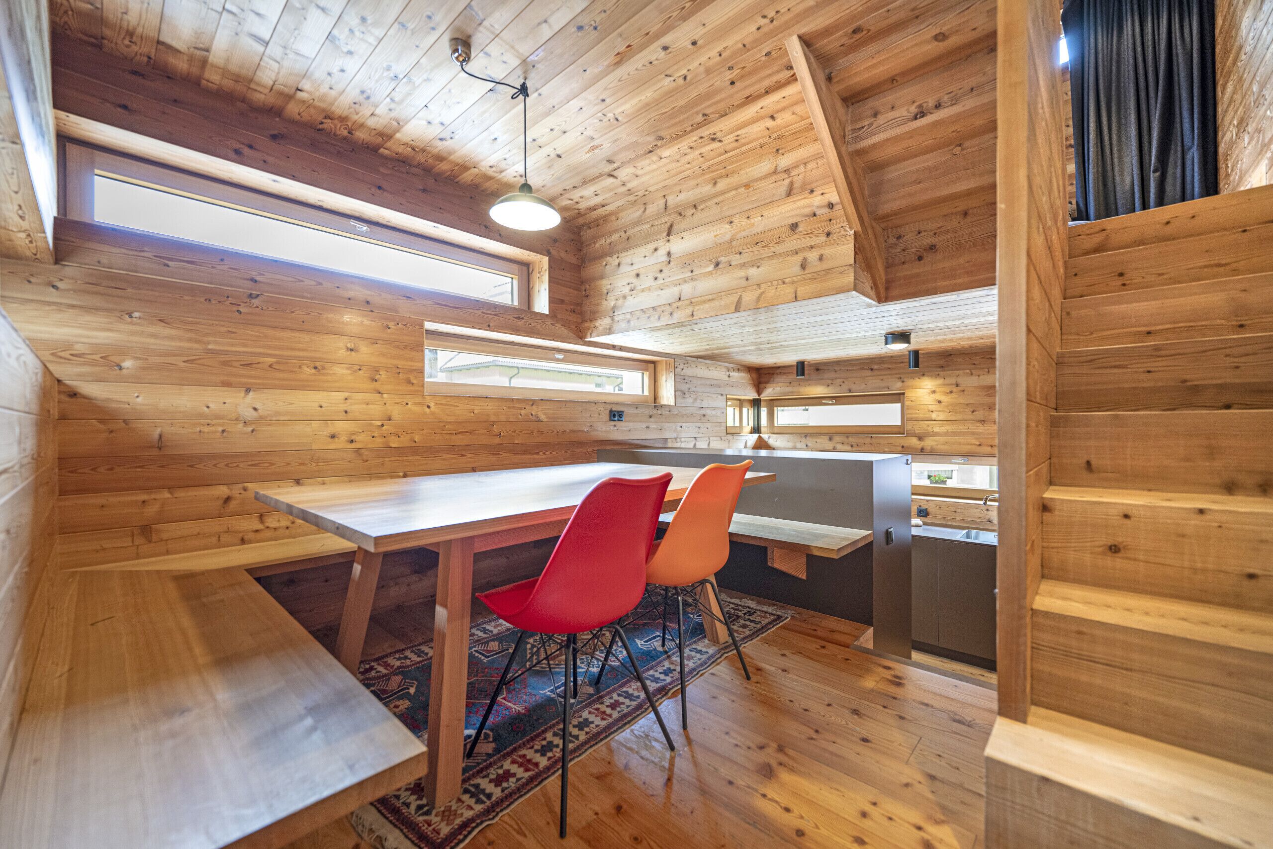A wooden interior with a dining table, two chairs, a rug, and a kitchen area.