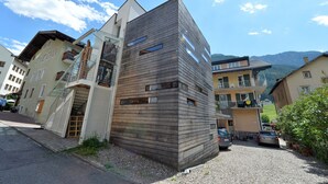 Um edifício moderno com fachada de madeira, uma escadaria que leva a uma varanda, e vista da área circundante, incluindo outros edifícios e montanhas.