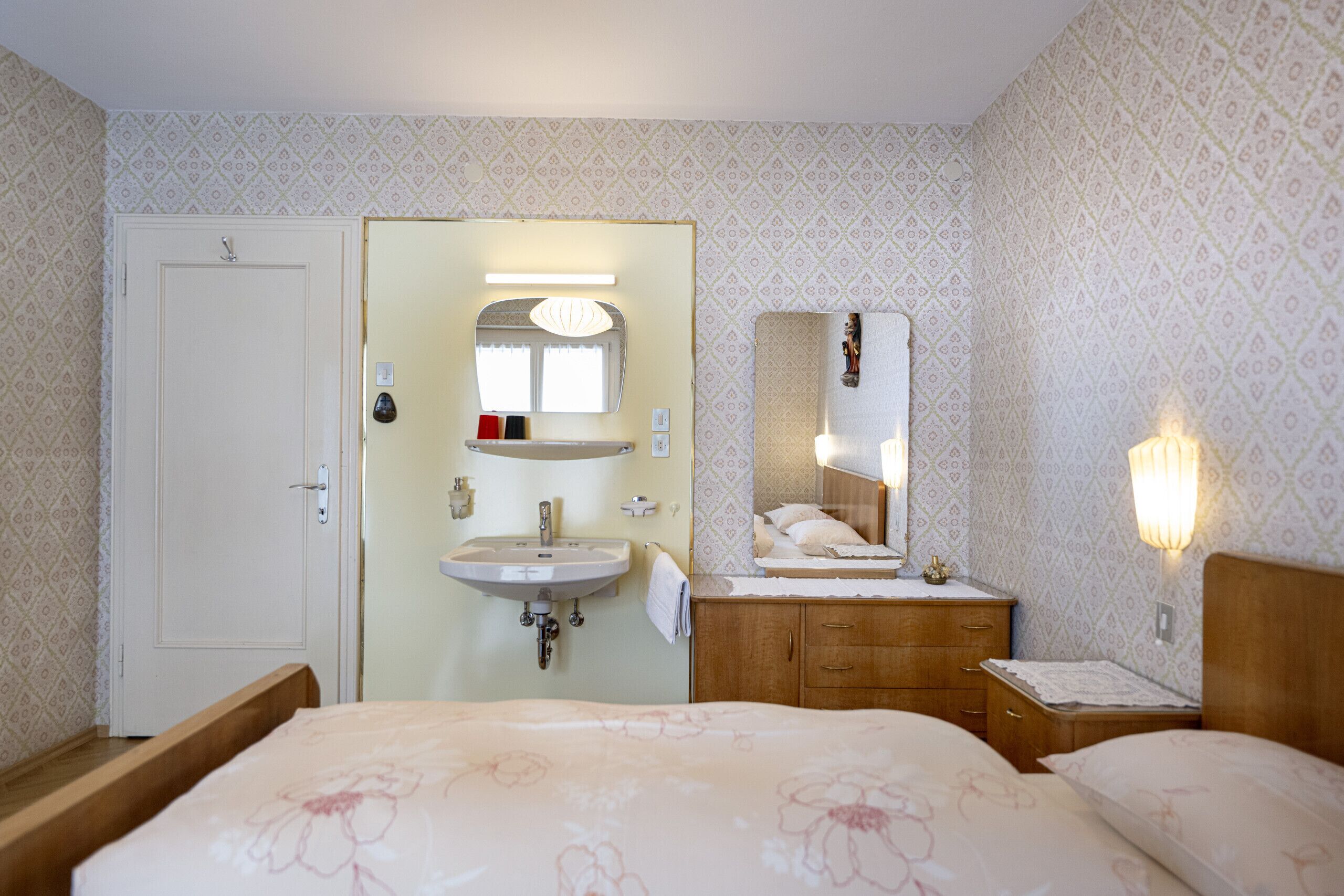 A hotel room with a bed, a bedside table, a nightstand, a mirror, a sink, and a door.