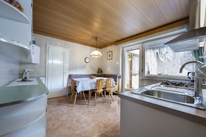 A compact kitchen with a dining area, wooden ceiling, and a window with curtains.