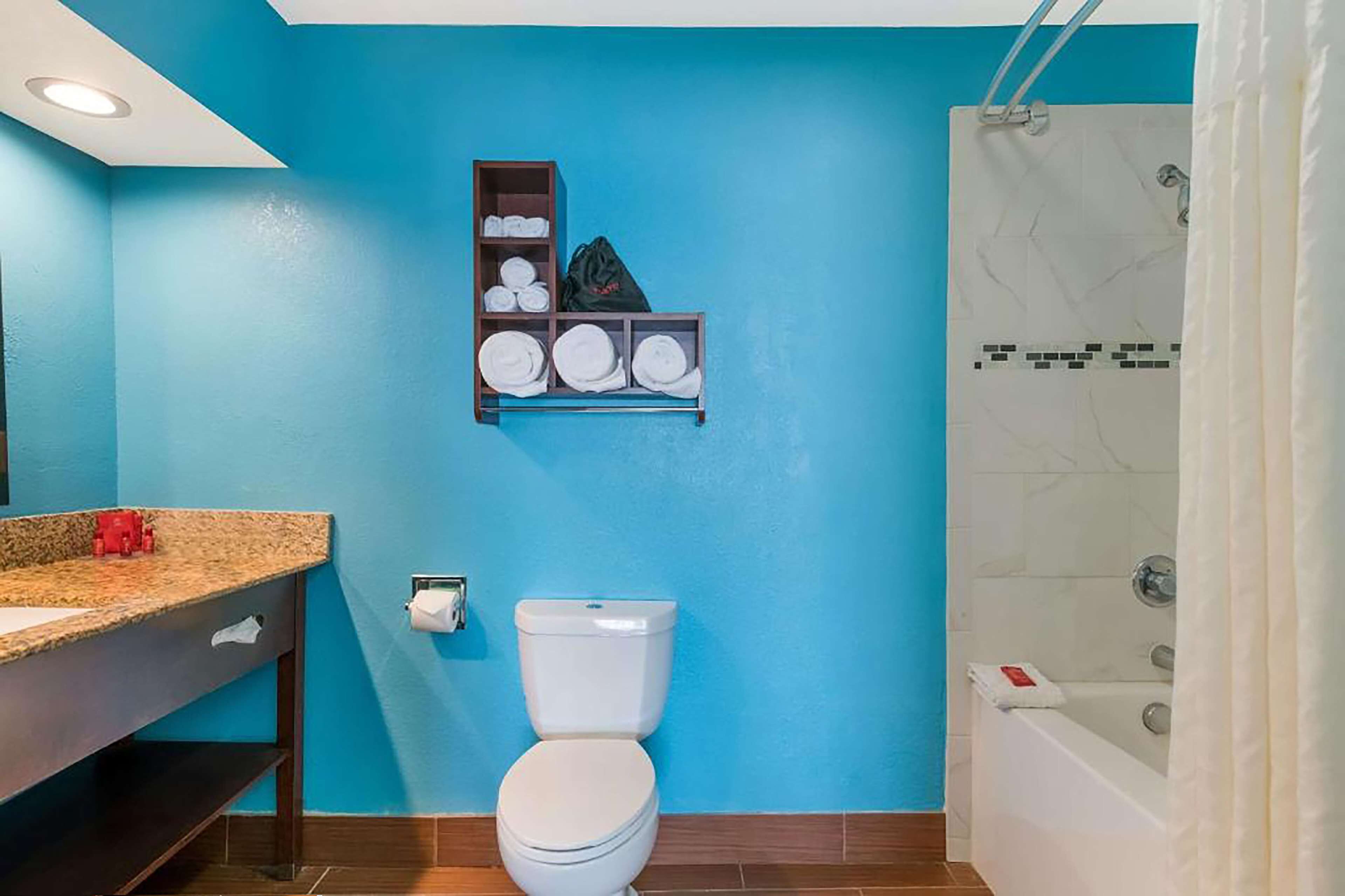 Une salle de bain avec un mur bleu, des toilettes blanches, une étagère avec des serviettes, un lavabo avec un plan de travail en granit et un espace douche.