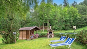 A wooden cabin with a small porch and a brick fireplace, surrounded by lush greenery and two striped lounge chairs. - Apartments Alara 134B (Ortisei)