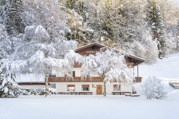 Un chalet enneigé, avec une façade en bois, une enseigne indiquant « GARNI » et une entrée bordée de bancs.