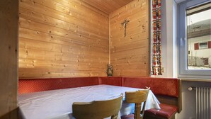 Een houten interieur met een tafel, stoelen, een bank, een kruisbeeld en een raam met uitzicht op een gebouw.