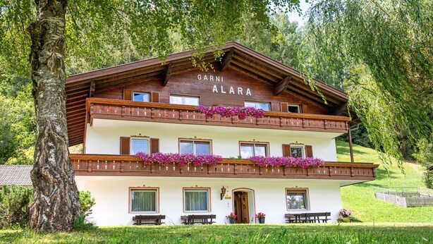 Een chalet met twee verdiepingen, genaamd "GARNI ALARA", met balkons, houten leuningen en bloembakken.