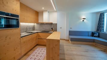 A modern kitchen with wooden cabinets, a built-in oven, and a grey countertop. There is a grey sofa with a patterned cushion in the living area.