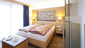 Una habitación de hotel moderna con una cama de madera, lámparas de noche, cabina de ducha de vidrio y un gran ventanal con cortinas.