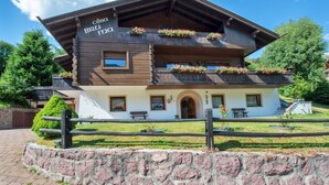 Ein traditionelles Holzchalet mit Balkonen, die mit Blumen geschmückt sind, einer Steinmauer im Vordergrund und blauem Himmel.