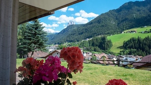 Uitzicht vanaf een balkon, met bloemen op de voorgrond, een bergketen op de achtergrond en een dorpje beneden.