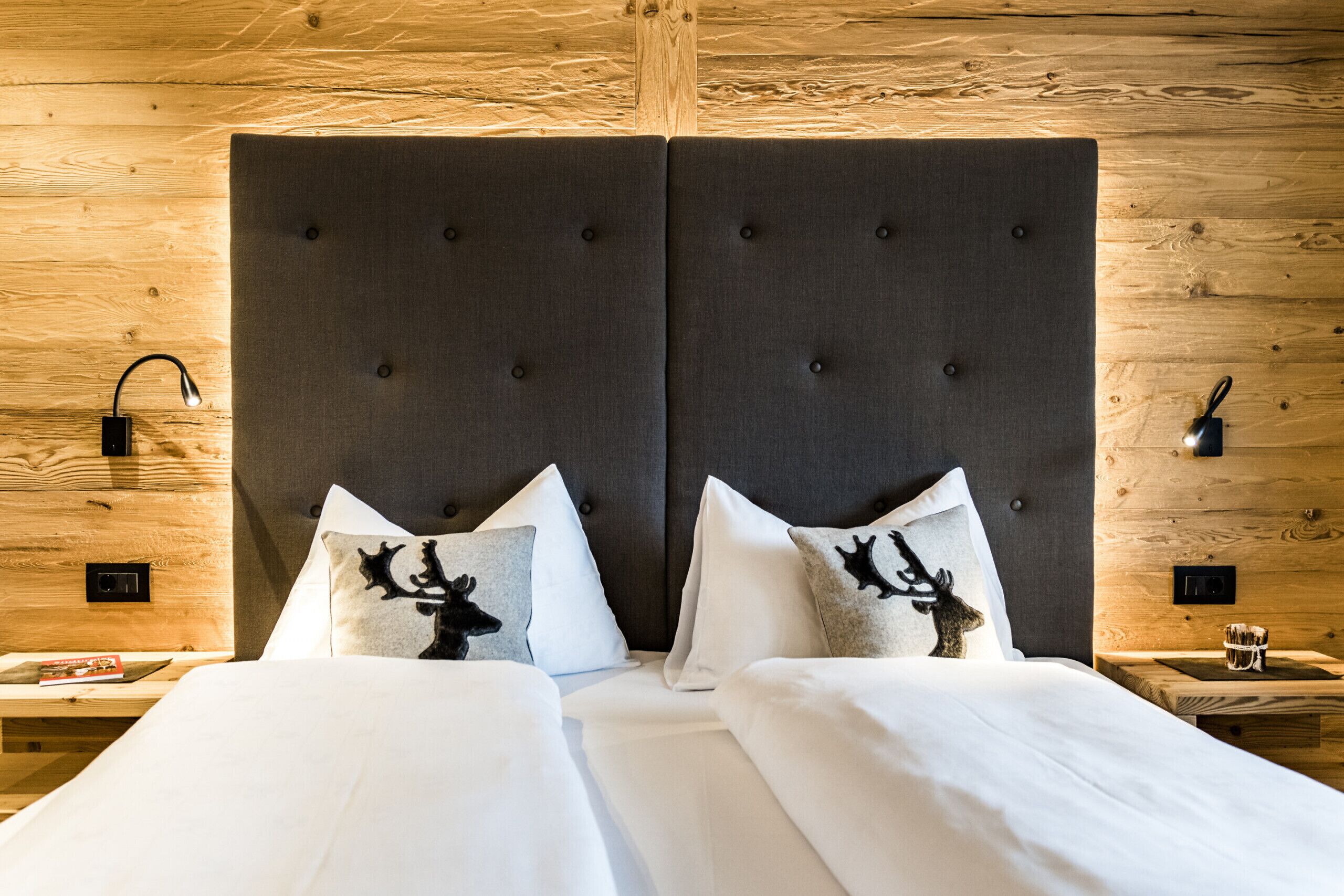 Una cama doble con sábanas blancas y dos almohadas decorativas con siluetas de ciervos. La habitación tiene un cabecero de madera y una mesita de noche con una lámpara y un vaso.