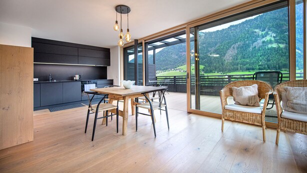 A modern kitchen with dark cabinets, a wooden dining table, and a large sliding glass door leading to a balcony with wicker furniture and a scenic mountain view. - Apartments Villa Mayr 125A (Ortisei)