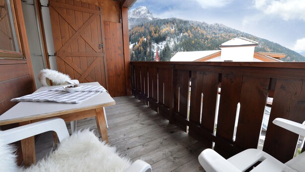 A wooden deck with a table and chairs, a white furry rug, and a view of a mountain with a church. - Apartments Nogler Frieda 124A (Selva di Val Gardena)